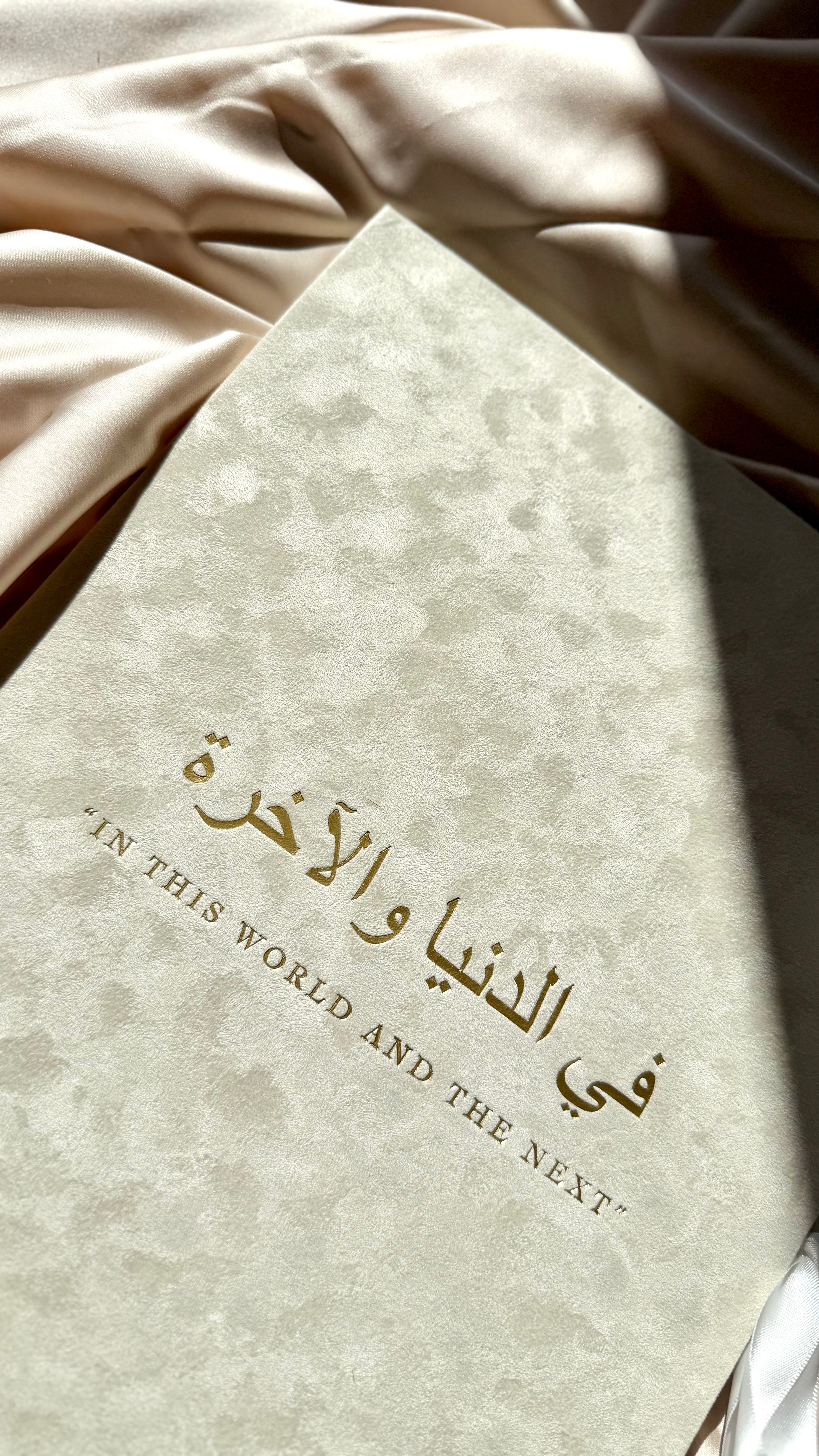 A close-up of a textured cream-colored book or notebook cover with elegant gold Arabic calligraphy and the English phrase "IN THIS WORLD AND THE NEXT" printed below it, resting on draped satin fabric.